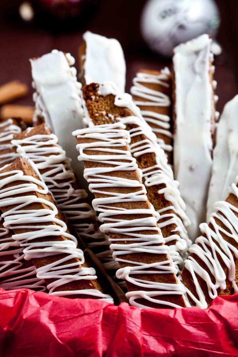 Close-up of gingerbread biscotti with white chocolate drizzle, arranged vertically in a red paper-lined container.