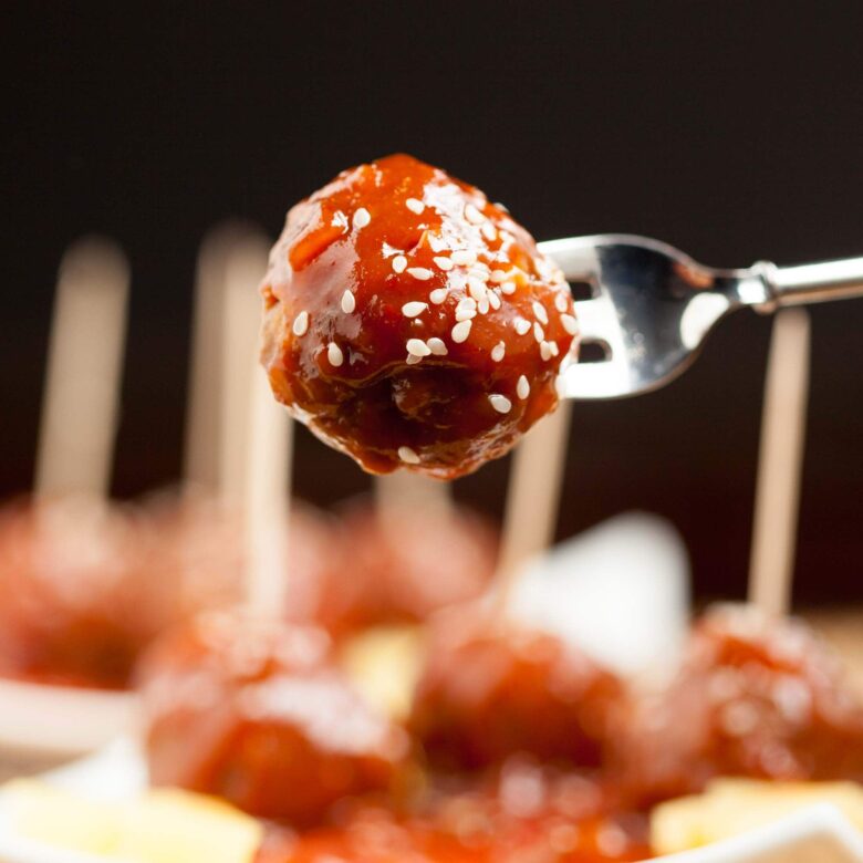 A fork holding a glazed sweet and sour meatball topped with sesame seeds, with more meatballs blurred in the background.