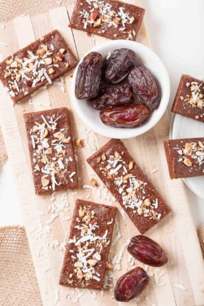 Rectangular date bars topped with shredded coconut and nuts are arranged on a wooden board.
