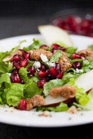 A plate of Pomegranate Salad featuring lettuce, pomegranate seeds, crumbled cheese, candied nuts, and slices of pear.