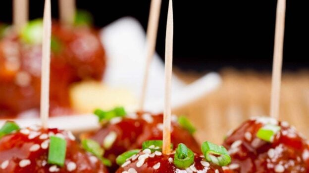 Close-up of sweet and sour meatballs on skewers, garnished with green onions and sesame seeds.