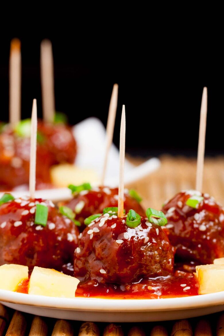 Close-up of sweet and sour meatballs on skewers, garnished with green onions and sesame seeds.
