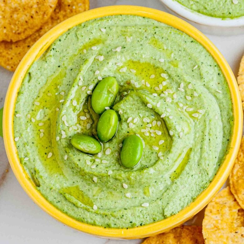 A bowl of green edamame hummus garnished with edamame beans and sesame seeds, surrounded by tortilla chips.