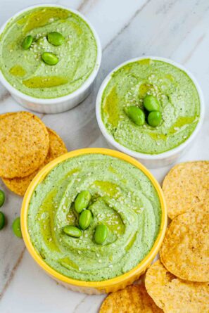 Three bowls of green edamame hummus topped with edamame beans and sesame seeds, surrounded by round tortilla chips on a marble surface.