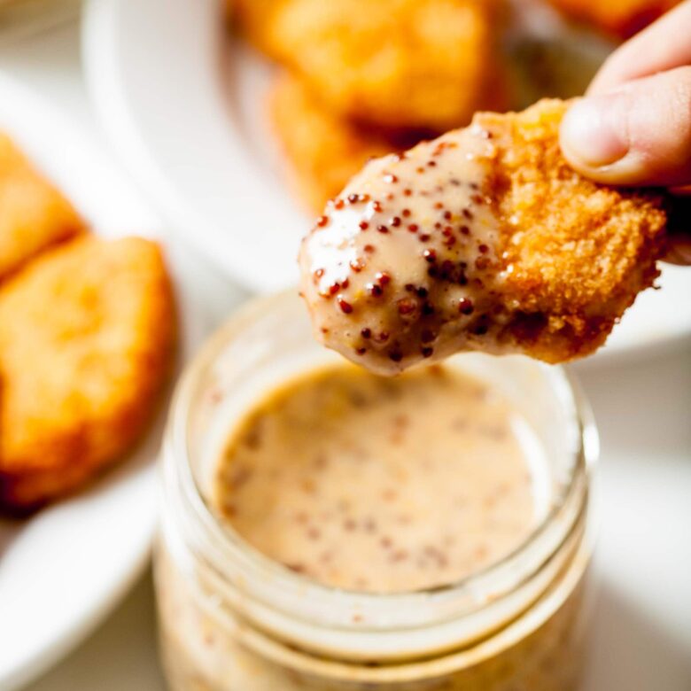 A hand dips a chicken nugget into a jar of grainy homemade honey mustard sauce.