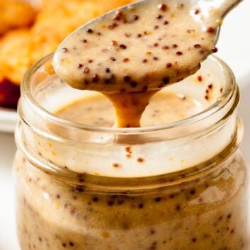Close-up shot of a spoon lifting creamy honey mustard sauce with visible seeds from a glass jar.