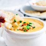Ranka panardina lustą į queso dubenį, ant kurio viršuje su pjaustytais žaliais svogūnais ir paprikomis.