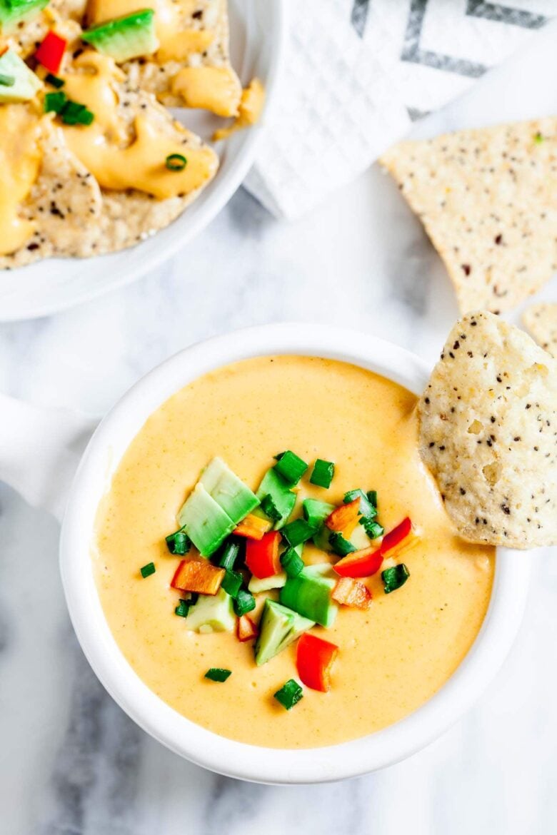 A bowl of queso dip topped with diced avocado, jalapeños, and red peppers, with a tortilla chip dipped in.