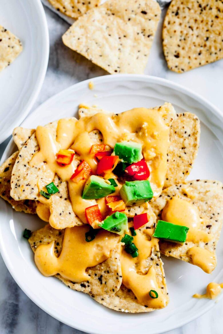 Nachos plokštelė su queso panardinimu, supjaustyta raudonaisiais paprikomis, žaliuoju avokadu ir supjaustytais žaliaisiais svogūnais.