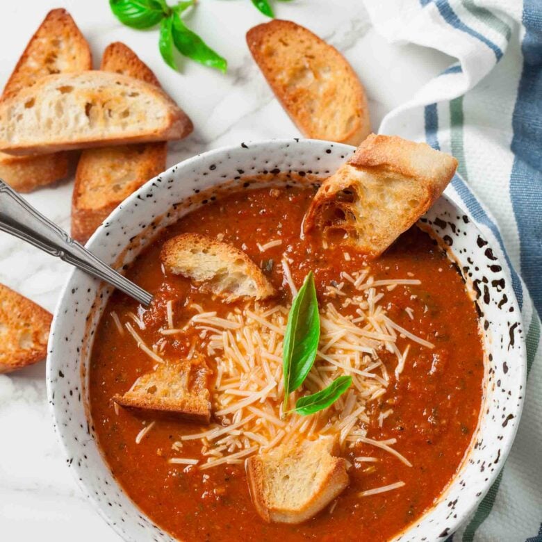 Overhead shot of bowl of roasted tomato soup topped with shredded cheese, croutons, and a basil leaves with slices of toasted bread on the side.