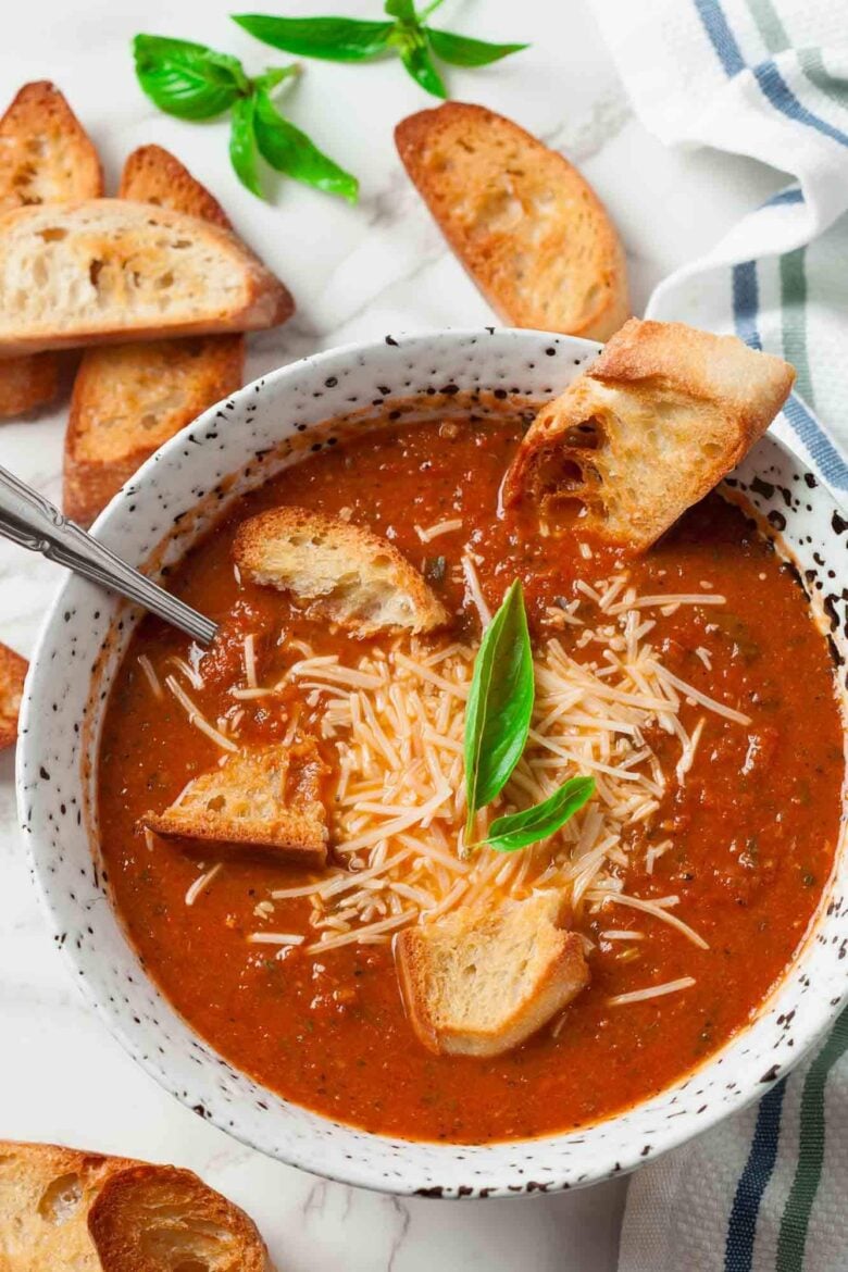 Overhead shot of bowl of roasted tomato soup topped with shredded cheese, croutons, and a basil leaves with slices of toasted bread on the side.