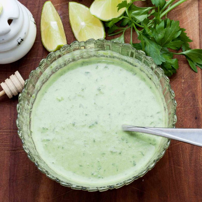Overhead shot fo a bowl of creamy green cilantro dressing with a spoon, surrounded by lime wedges and fresh parsley on a wooden surface.