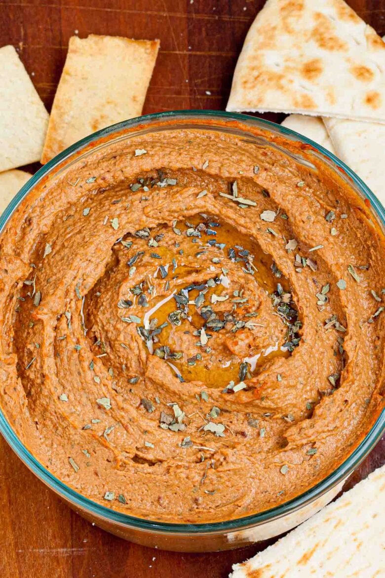 A bowl of sun dried tomato hummus garnished with herbs and olive oil, surrounded by pita bread and crackers on a wooden surface.