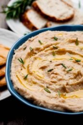 Bowl of white bean dip garnished with rosemary, with sliced bread and crackers in the background.