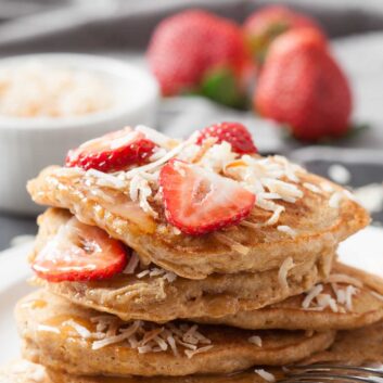 A stack of fluffy coconut pancakes topped with sliced strawberries and shredded coconut, served on a white plate with a fork.