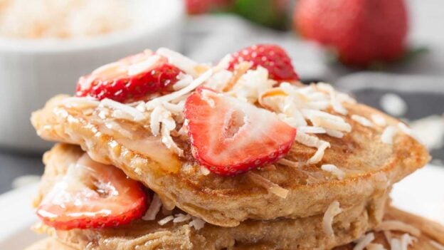 A stack of fluffy coconut pancakes topped with sliced strawberries and shredded coconut, served on a white plate with a fork.