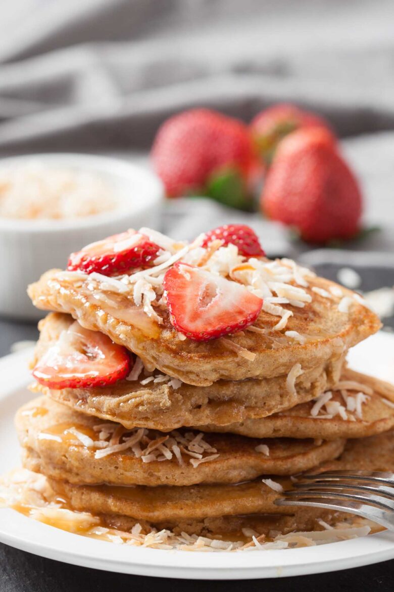 A stack of fluffy coconut pancakes topped with sliced strawberries and shredded coconut, served on a white plate with a fork.