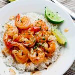 Overhead shot of a plate of rice topped with Honey Lime Shrimp in sauce, garnished with herbs and lime wedges.