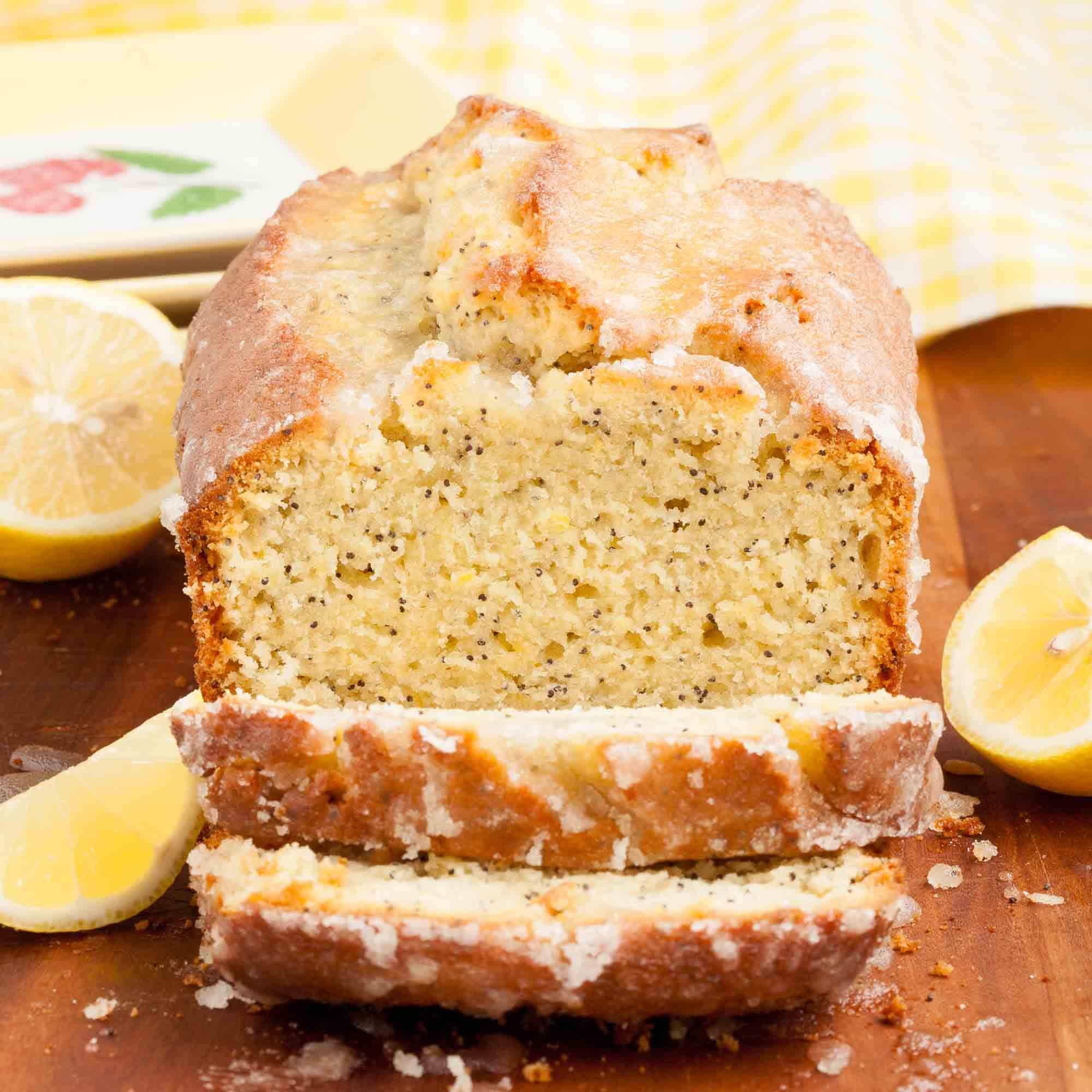 Sliced lemon poppy seed bread with a glazed topping, on a wooden surface with lemon halves on the side.