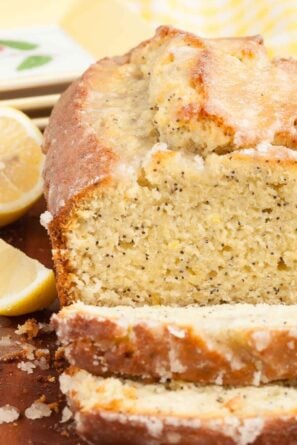 Sliced lemon poppy seed bread with a glazed topping, on a wooden surface with lemon halves on the side.