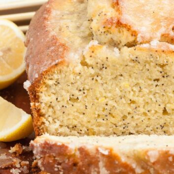 Sliced lemon poppy seed bread with a glazed topping, on a wooden surface with lemon halves on the side.
