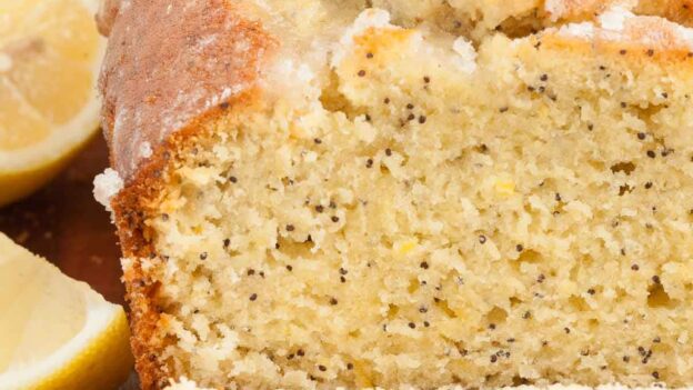 Sliced lemon poppy seed bread with a glazed topping, on a wooden surface with lemon halves on the side.