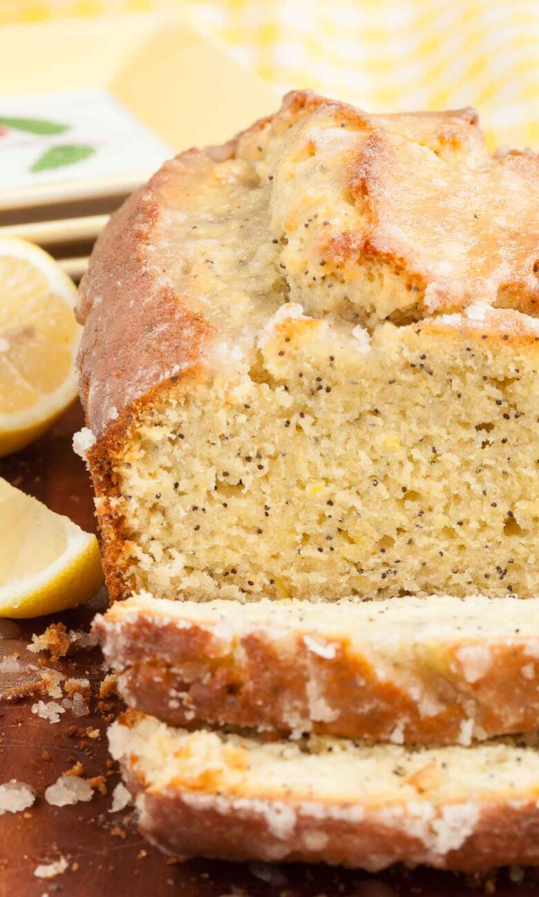Sliced lemon poppy seed bread with a glazed topping, on a wooden surface with lemon halves on the side.
