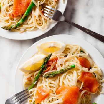 Two plates of lemon pasta pasta with smoked salmon, asparagus, lemon wedges, and forks on a marble surface.