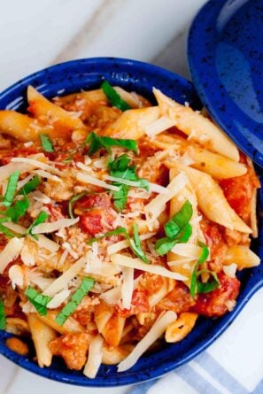 A blue bowl filled with penne with vodka sauce topped with shredded cheese, and garnished with fresh basil leaves.