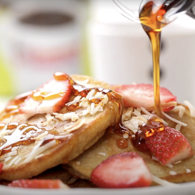 Pancake syrup being poured over freshly made coconut pancakes, with shredded coconut and chopped strawberries on top.