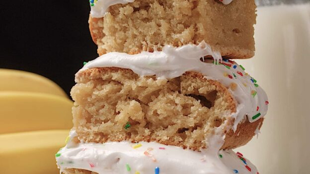 Three slices of banana cake with cream cheese frosting and sprinkles on top stacked on a plate.