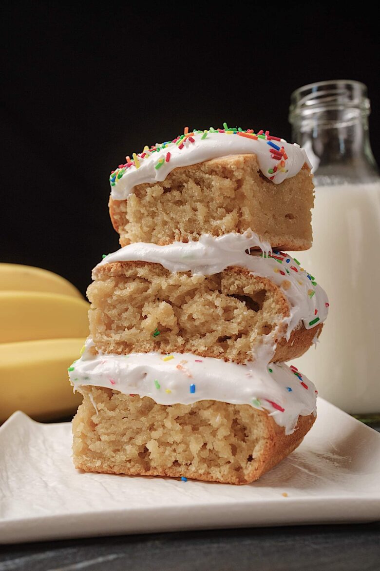Three slices of banana cake with cream cheese frosting and sprinkles on top stacked on a plate.