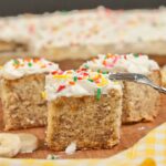 Slices of cream cheese frosted banana cake topped with colorful sprinkles on a wooden board.