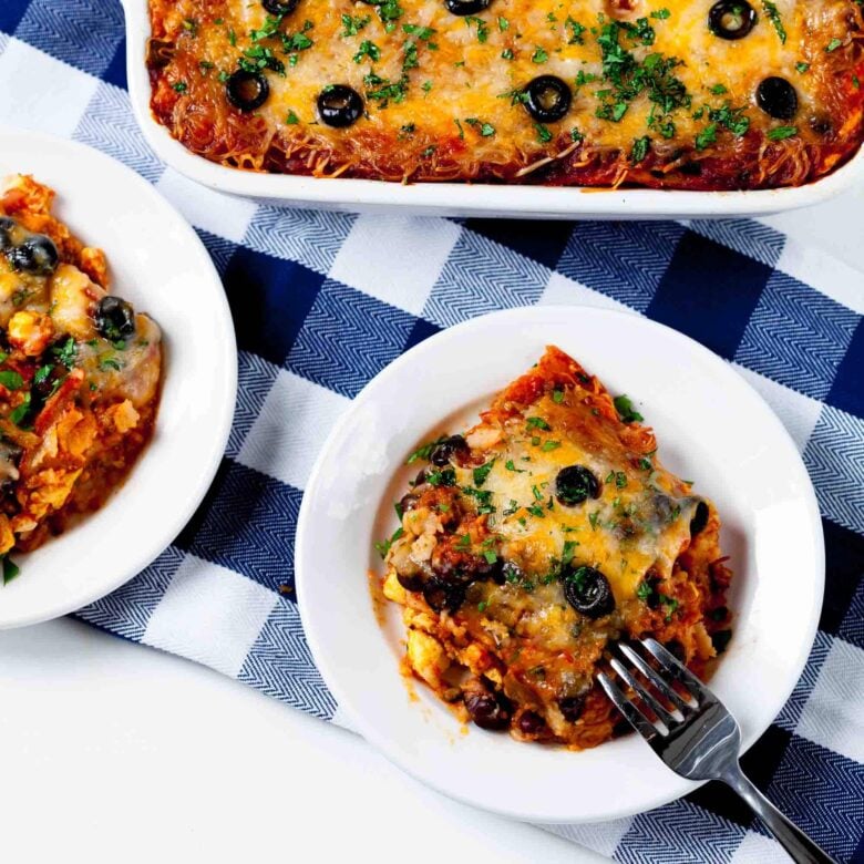 Baked Mexican breakfast casserole with melted cheese and black olives on a blue checkered cloth. Two servings on white plates with forks.