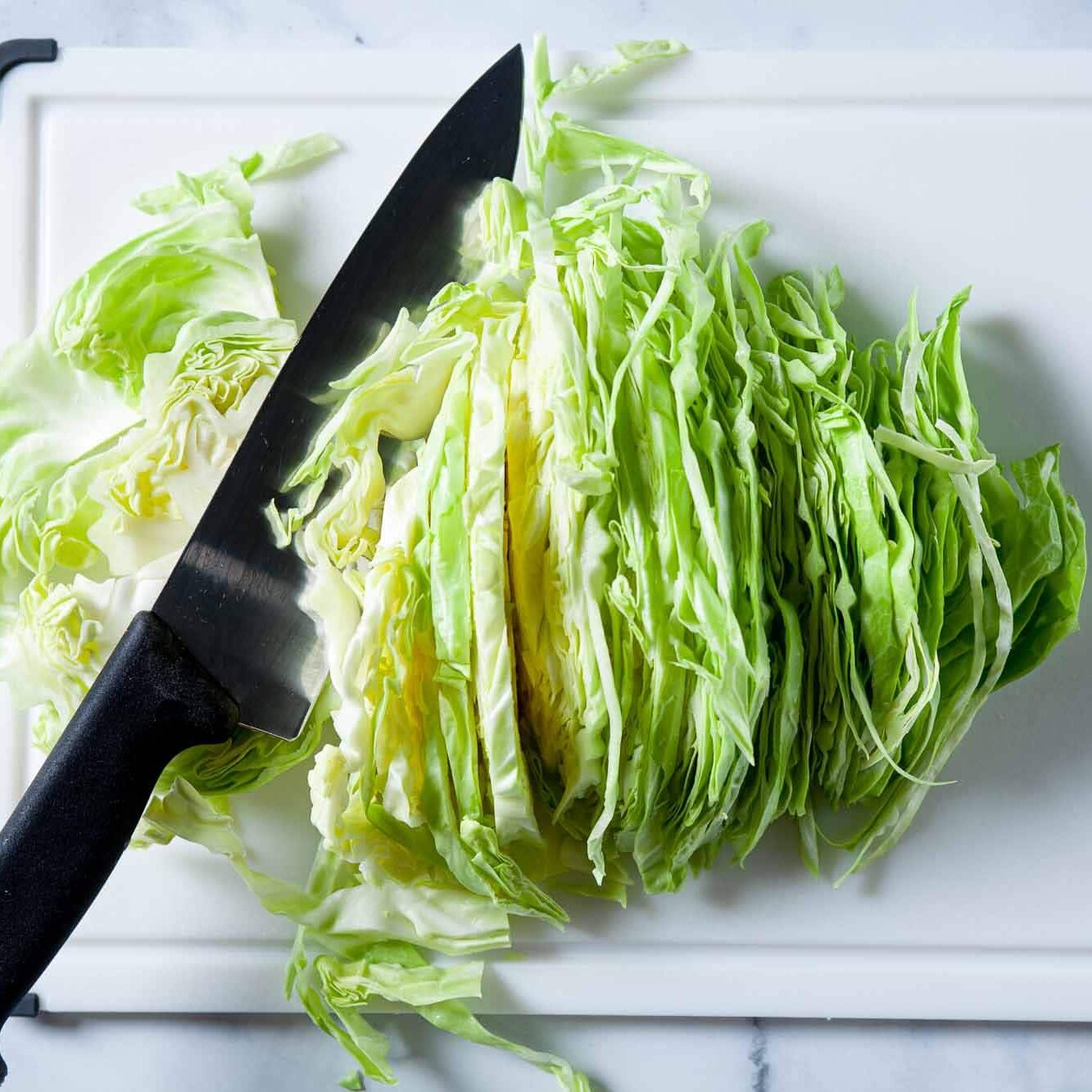 Finely sliced green cabbage with a chef's knife on a white chopping board.