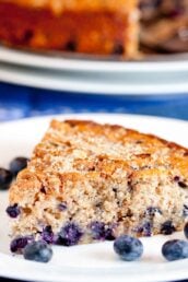 A close-up of a moist blueberry coffee cake slice on a white plate, with juicy blueberries baked inside and a few fresh blueberries nearby.