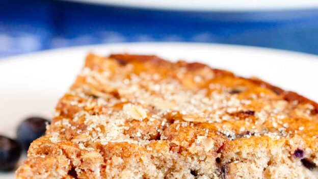 A close-up of a moist blueberry coffee cake slice on a white plate, with juicy blueberries baked inside and a few fresh blueberries nearby.