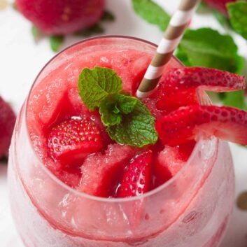 A glass of strawberry frosé made using a classic frosé recipe, topped with fresh strawberry slices and mint leaves, served with a striped straw.