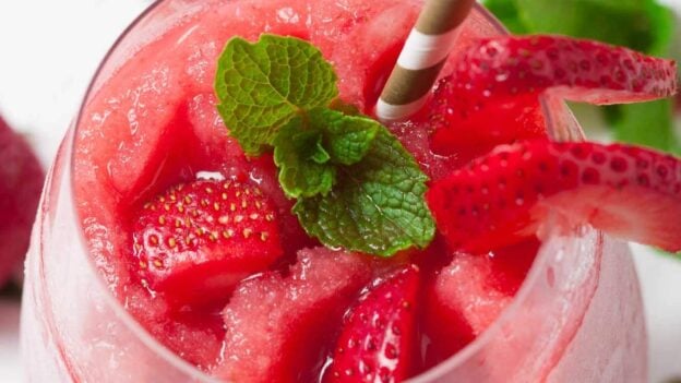 A glass of strawberry frosé made using a classic frosé recipe, topped with fresh strawberry slices and mint leaves, served with a striped straw.