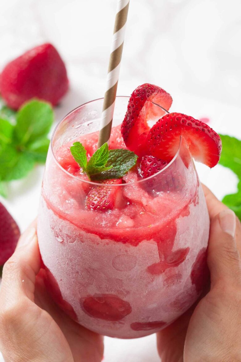 A hand holds a frosty glass filled with a strawberry slushie, made using an easy Frosé recipe, garnished with fresh strawberry slices and a mint sprig. A striped paper straw is in the drink.