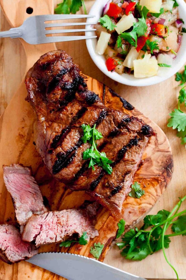 Grilled marinated steak with perfect grill marks and parsley garnish on a wooden board, two sliced pieces in front, served with a bowl of pineapple salsa to assembled grilled steak tacos.