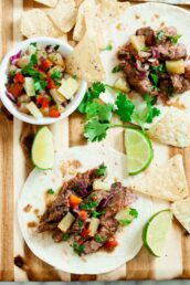 Two Grilled Steak Tacos with pineapple salsa sit on a wooden board, surrounded by tortilla chips, lime wedges, and fresh cilantro.