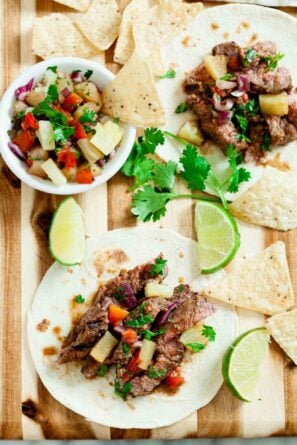 Two Grilled Steak Tacos with pineapple salsa sit on a wooden board, surrounded by tortilla chips, lime wedges, and fresh cilantro.