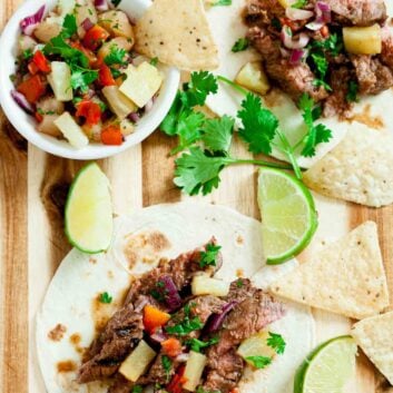 Two Grilled Steak Tacos with pineapple salsa sit on a wooden board, surrounded by tortilla chips, lime wedges, and fresh cilantro.