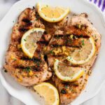 Overhead shot of grilled lemon chicken with sliced lemon wedges on top and chopped herbs.