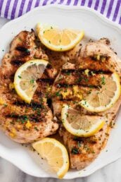 Overhead shot of grilled lemon chicken with sliced lemon wedges on top and chopped herbs.