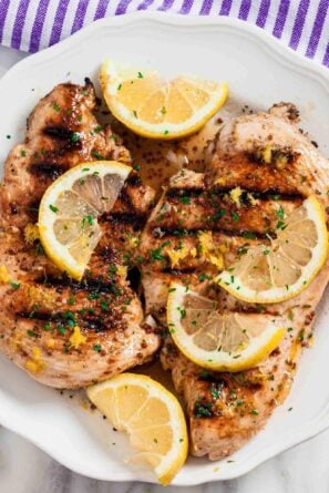 Overhead shot of grilled lemon chicken with sliced lemon wedges on top and chopped herbs.