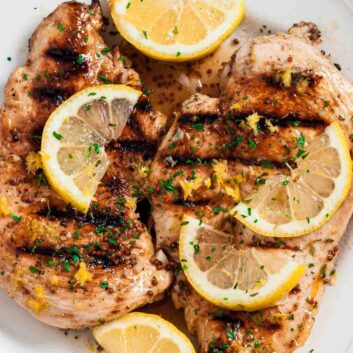 Overhead shot of grilled lemon chicken with sliced lemon wedges on top and chopped herbs.