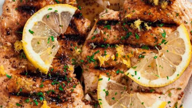 Overhead shot of grilled lemon chicken with sliced lemon wedges on top and chopped herbs.