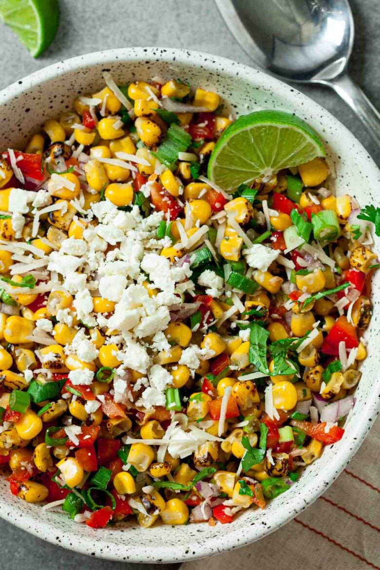 A quick and flavorful Mexican Street Corn Salad, this bowl features crumbled cheese, chopped herbs, red bell peppers, and a lime wedge, with a spoon nearby for easy serving.
